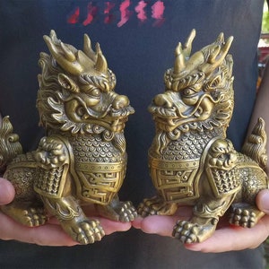 May include: Two bronze-colored, mythical creature statues. Each statue has a detailed design, including scales, horns, and a fierce expression. The statues are held in front of a dark background.