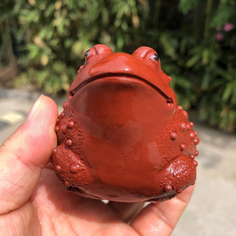 May include: A reddish-brown frog figurine with a textured surface. The frog has a wide mouth and small, black eyes. The figurine is held in a hand, with a blurred green background.