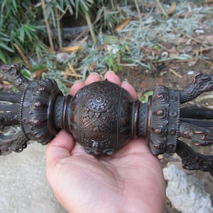 May include: A bronze-colored vajra, a ritual object with ornate detailing. The central sphere features intricate patterns, flanked by dragon-like figures. The object is held in a hand, showcasing its size and craftsmanship.