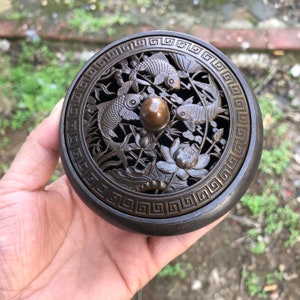 Peut inclure: Un brûleur d'encens rond de couleur bronze avec un couvercle détaillé représentant des carpes koï sculptées, des fleurs de lotus et du feuillage. Le couvercle a un bouton central et une bordure décorative. Le brûleur est tenu dans une main.