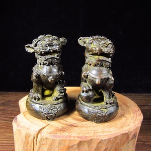 May include: Two bronze-coloured Foo Dog statues. Each statue is in a seated position with detailed features, including a mane, collar, and paws resting on a ball. The statues are displayed on a wooden base with a natural wood grain.