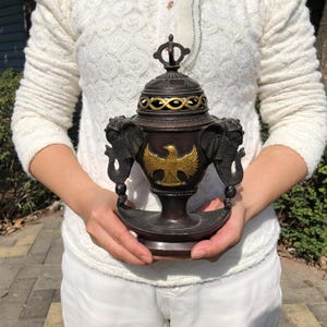 May include: A bronze-colored incense burner with a decorative lid and a gold eagle emblem. The burner features elephant head handles and intricate gold accents. The base is round and the overall design is ornate.