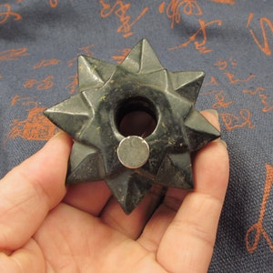 Puede incluir: Un objeto en forma de estrella de color verde oscuro con un agujero central y bordes puntiagudos. El objeto parece estar hecho de piedra o un material similar. Un pequeño objeto redondo de color plateado se coloca en la superficie de la estrella.