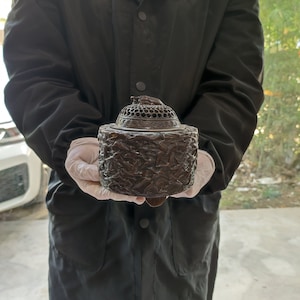 Op de afbeelding: Een donkerbruine, cilindrische wierookbrander met een gedetailleerd, gebeeldhouwd vogelontwerp. Het deksel is geperforeerd met een decoratief patroon en bekroond met een kleine vogel. De brander wordt vastgehouden door een persoon die een zwarte jas en witte handschoenen draagt.