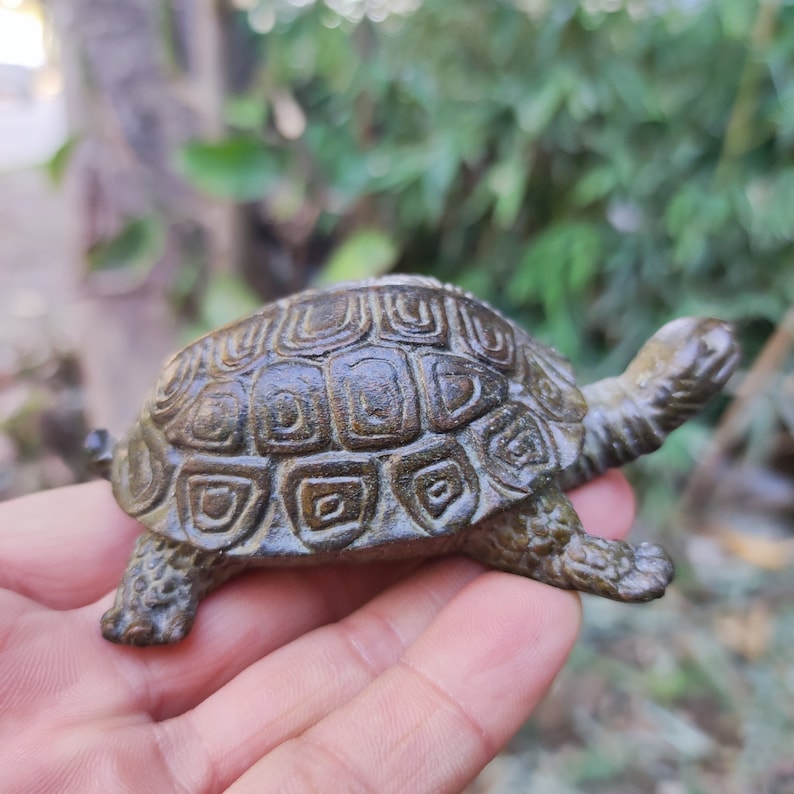 May include: A small, bronze-colored turtle figurine with a detailed shell pattern. The sculpture is held in a hand, showcasing its size and intricate design. The turtle's head and legs are visible, adding to its realistic appearance.