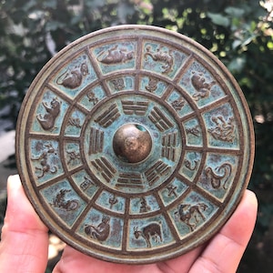 Op de afbeelding: Een ronde, bronskleurige decoratieve plaat met een centrale knop. De plaat heeft een cirkelvormig ontwerp met dierenfiguren en Chinese karakters. De plaat heeft een verweerde, antieke uitstraling met hints van blauwgroene patina.