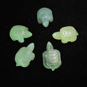Könnte beinhalten: Fünf durchscheinende grüne Schildkrötenfiguren auf einer schwarzen Oberfläche. Jede Schildkröte hat einen detaillierten Panzer, einen kleinen Kopf und Beine. Die Schildkröten variieren leicht in der Farbe, von hell- bis mittelgrün.