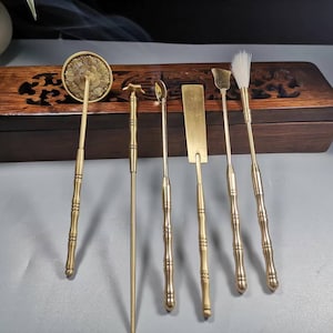 Könnte beinhalten: Ein Set aus sieben Teezeremonie-Werkzeugen aus Messing. Die Werkzeuge umfassen einen runden Schöpfer, eine kleine Gabel, einen Löffel, einen flachen Spatel, eine kleine Schaufel und einen Pinsel. Die Werkzeuge haben dekorative Griffe.