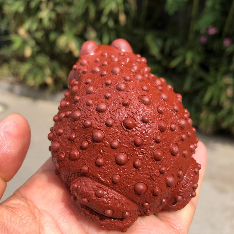 May include: A reddish-brown frog figurine with a textured, bumpy surface. The frog is held in a hand, showcasing its detailed design. The figurine's color and texture give it a unique, handcrafted appearance.