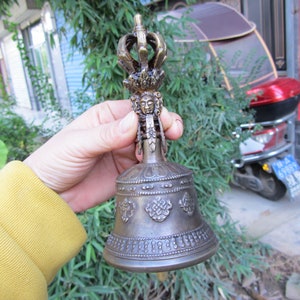 May include: Bronze-colored bell with intricate detailing. The bell features a crown-like top, a detailed face, and ornate patterns. The bell is held in a hand, with a green background.