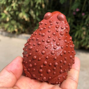 May include: A reddish-brown frog figurine with a textured surface covered in small, round bumps. The frog has a rounded body and is held in a person's hand. The background is blurred with green foliage.