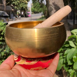 May include: A golden singing bowl with a wooden striker rests on a red and gold cushion. The bowl has a smooth, polished surface and a simple, elegant design. The wooden striker is light-colored and cylindrical.