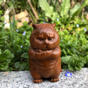 Può includere: Piccola statuetta in legno intagliato raffigurante un gatto con un'espressione accigliata. Il gatto marrone, con le braccia incrociate, presenta dettagli della venatura del legno. La statuetta è su una superficie di pietra, con sfondo di vegetazione.