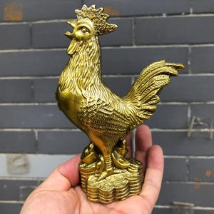 May include: A golden rooster figurine with detailed feather textures and a prominent comb. The rooster stands atop a base of stacked coins, suggesting prosperity. The statue is made of a shiny, metallic material.
