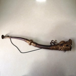 May include: A decorative horn with a dragon head design. The horn is primarily brown with gold-colored accents, including ornate detailing around the body and the dragon's head. A chain is attached to the horn.