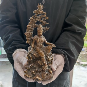 May include: A bronze-colored statue of a seated figure, possibly a deity, with ornate details. The figure is adorned with jewelry and seated on a rocky base with a stylized tree or cloud formation behind. The statue is held by gloved hands.