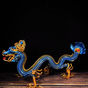Könnte beinhalten: Detaillierte Drahtskulptur eines Drachen, hauptsächlich blau mit goldenen Akzenten. Der Drache hat rote Perlen als Akzente und steht auf einer Holzoberfläche. Der Hintergrund ist schwarz. Die Skulptur zeigt filigrane Drahtarbeit.