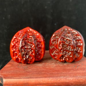 Puede incluir: Dos nueces de madera roja talladas con detalles intrincados. Las nueces se exhiben sobre una superficie de madera marrón rojiza sobre un fondo negro. Las tallas tienen un acabado brillante.