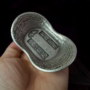 May include: A silver-colored metal ingot shaped like a traditional Chinese sycee, featuring a textured, rippled surface and a central rectangular panel with engraved Chinese characters including the text 縣解, 民國年月, and 中立錢局.