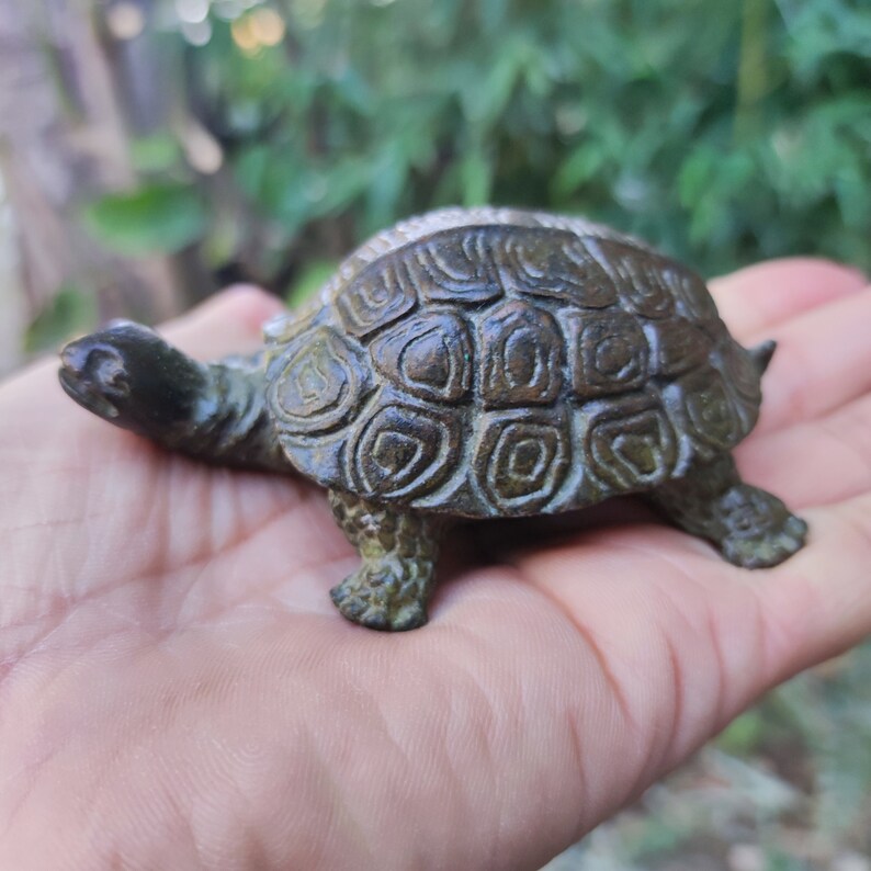 May include: A small, bronze-colored turtle figurine with detailed shell patterns. The turtle has a textured appearance and is posed with its head and legs extended. The figurine is likely a decorative item or collectible.