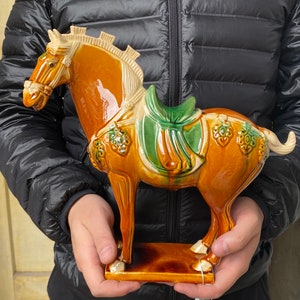 Puede incluir: Figura de caballo de cerámica con una silla de montar verde. El caballo es principalmente marrón con una melena y cola color crema. La figura es sostenida por una persona.