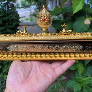 May include: Ornate gold-colored incense burner with a decorative design. The burner features two deer figurines and a central finial with a red stone. The base is a dark brown color with gold filigree.