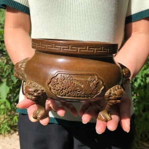 May include: Bronze-colored incense burner with intricate relief carvings. The vessel features a decorative band around the rim and detailed lion-head feet. The body of the burner showcases a detailed relief of a mythical bird.