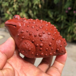 May include: A reddish-brown ceramic frog figurine with a textured, bumpy surface. The frog is in a seated position, with its eyes closed and mouth slightly open. The figurine is held in a person's hand.