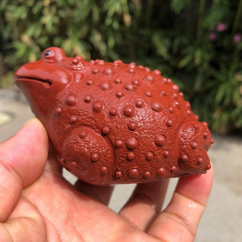 May include: A reddish-brown ceramic frog figurine with a textured, bumpy surface. The frog is in a seated position, with its eyes closed and mouth slightly open. The figurine is held in a person's hand.