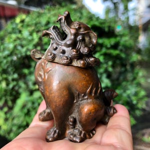 May include: A bronze-colored, detailed figurine of a mythical creature, possibly a guardian lion. The sculpture has an open mouth, intricate mane, and detailed paws. The figurine is held in a hand against a blurred green background.
