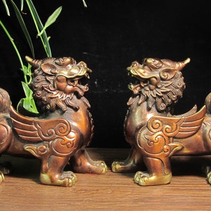 May include: Two bronze-colored mythical creatures with wings and open mouths. The statues have detailed features, including ornate wings and curled tails. They are positioned facing each other on a wooden surface, with greenery in the background.