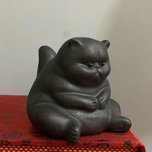 May include: A dark gray ceramic cat figurine in a seated position. The cat has a round body, small ears, and a slightly grumpy expression. Its paws are clasped together, and its tail is raised. The figurine is sitting on a red surface.