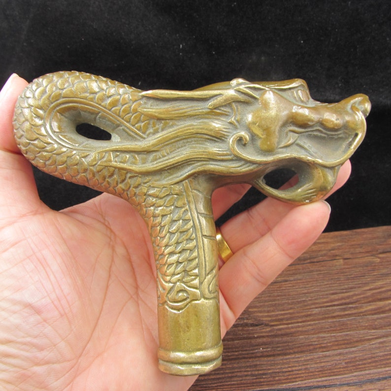 May include: A detailed bronze dragon head cane handle. The handle features a dragon's head with intricate scales and flowing mane. The handle is a warm, antique gold color.