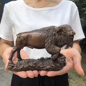 以下が含まれることがあります： 歩行ポーズのバイソンのブロンズ彫刻。彫刻は、ふさふさのたてがみとあごひげを備えた、詳細なテクスチャを持っています。バイソンはテクスチャのあるベースに取り付けられており、全体的な色は濃いブロンズです。