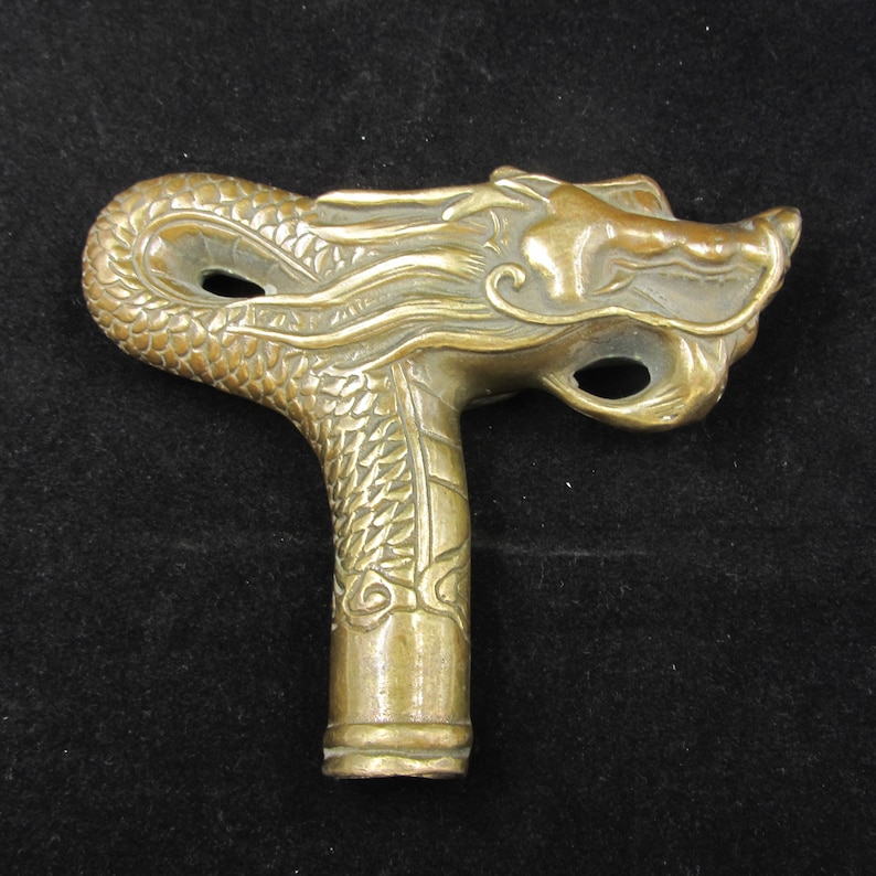 May include: A bronze-colored dragon head cane handle. The handle features intricate details of a dragon's face and scales. The handle is designed to be held in the hand and has a curved shape.