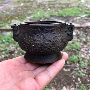 May include: A small, dark bronze incense burner with ornate floral and dragon detailing. The vessel has two dragon-shaped handles and sits on a small, round base. The exterior is covered in intricate relief patterns.