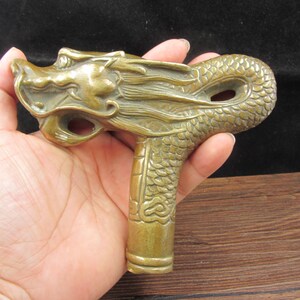 May include: A bronze-colored dragon head cane handle. The handle is intricately detailed with scales and a dragon's face. The handle is held in a hand, showcasing the detailed craftsmanship of the dragon design.