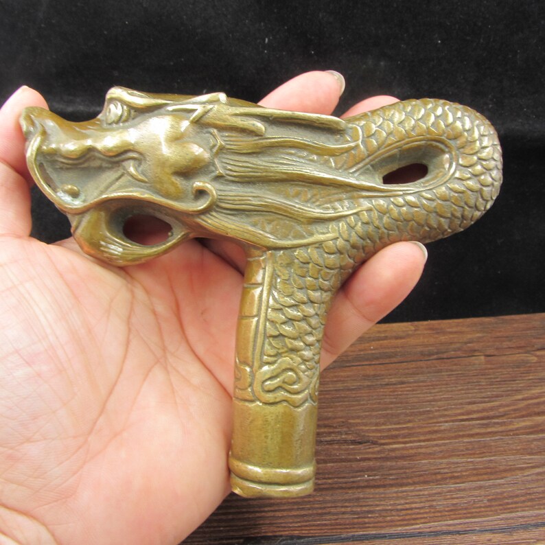 May include: A bronze-colored dragon head cane handle. The handle is intricately detailed with scales and a dragon's face. The handle is held in a hand, showcasing the detailed craftsmanship of the dragon design.