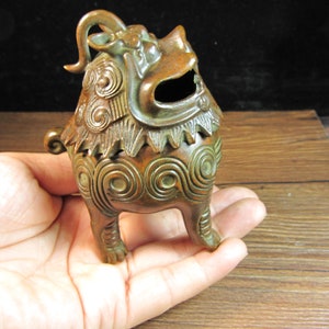 Può includere: Bruciatore di incenso color bronzo a forma di creatura mitica. Il bruciatore presenta intricati motivi a spirale e lineari, con una testa dettagliata e una bocca aperta. L'oggetto è tenuto in mano, mostrando le sue dimensioni e la sua maestria.