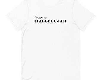 May include: White t-shirt with the phrase "raise a HALLELUJAH" printed in black. The word "raise a" is in a cursive font, while "HALLELUJAH" is in a bold, block font. The shirt is made of a soft material.
