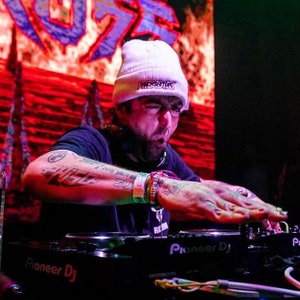 May include: A man with a white beanie with the word "RESPECT" embroidered on it is DJing with a Pioneer DJ mixer. He has tattoos on his arms and is wearing a black t-shirt.