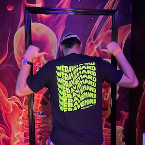 May include: A black t-shirt with a repeating pattern of the words "We Rave Hard" in neon green. The shirt is being worn by a person in front of a wall with a fiery skeleton design.