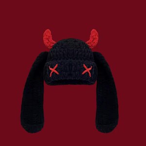 May include: A black crocheted beanie featuring two red horns on top and two long, dangling side flaps. The front of the beanie displays two red embroidered X shapes, one on each side, positioned where eyes would typically be on a face.