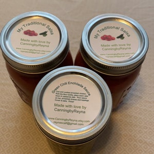 May include: Three glass jars with red sauce. The labels on the jars read "My Traditional Salsa" and "Green Chili Enchilada Sauce". Each label includes the text "Made with love by CanningbyReyna" and contact information for the company.