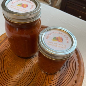 May include: Two glass jars filled with a red sauce, labeled "Mango Habanero Salsa" with the text "Made with love by CanningbyReyna". The jars are sitting on a wooden tray.