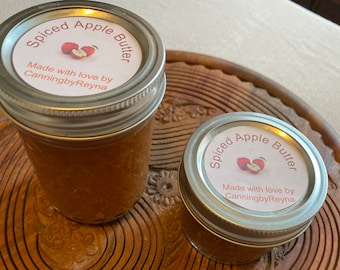 May include: Two glass jars of spiced apple butter with white labels that say "Spiced Apple Butter" and "Made with love by CanningbyReyna". The jars are sitting on a wooden surface.