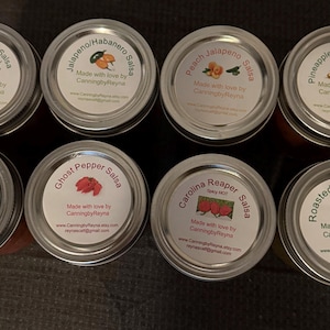 May include: Ten glass jars with silver lids, each containing a different type of salsa. The jars are labeled with the type of salsa, including "My Traditional Salsa", "Jalapeno/Habanero Salsa", "Peach Jalapeno Salsa", "Pineapple Jalapeno Salsa", "Mango Habanero Salsa", "Ghost Pepper Salsa", "Carolina Reaper Salsa", and "Roasted Salsa Verde". Each label includes the text "Made with love by CanningbyReyna".