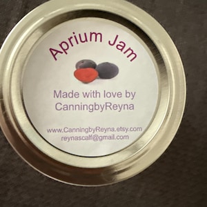 May include: A jar lid with a label that reads "Aprium Jam" in purple script. The label features illustrations of a red and two dark purple fruits. The text "Made with love by CanningbyReyna" is also visible.