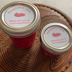 May include: Two jars of raspberry jam with silver lids sit on a wooden serving tray. The labels read "Raspberry Jam" with a raspberry illustration and "Made with love by CanningbyReyna."
