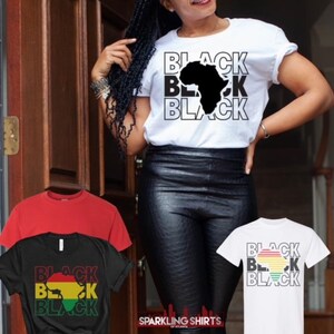 May include: White t-shirt with the word "BLACK" stacked three times, with a black silhouette of the African continent. Additional t-shirts in red and black are also visible, each with variations of the same design. The shirts are from Sparkling Shirts of Atlanta.